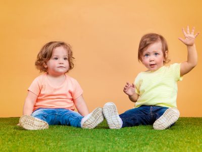 Happy funny girl twins sisters sitting on grass and playing and laughing on orange background